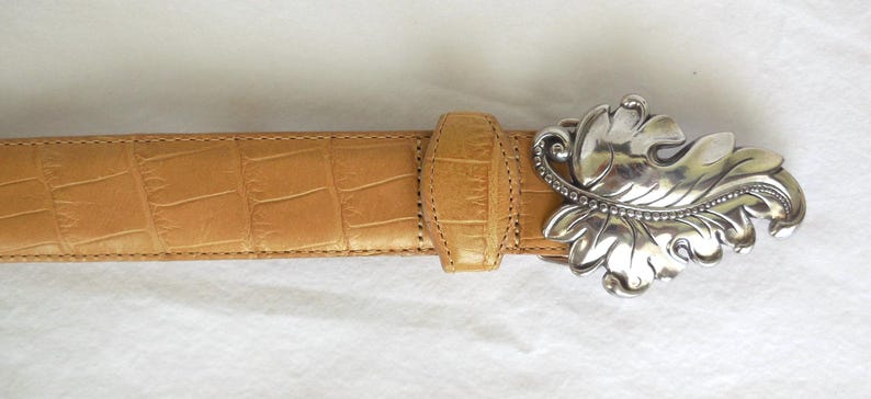 May include: Tan leather belt with a silver-coloured, ornate leaf-shaped buckle. The belt has a crocodile skin texture. The buckle is detailed with small, round embellishments. A stylish accessory for any outfit.