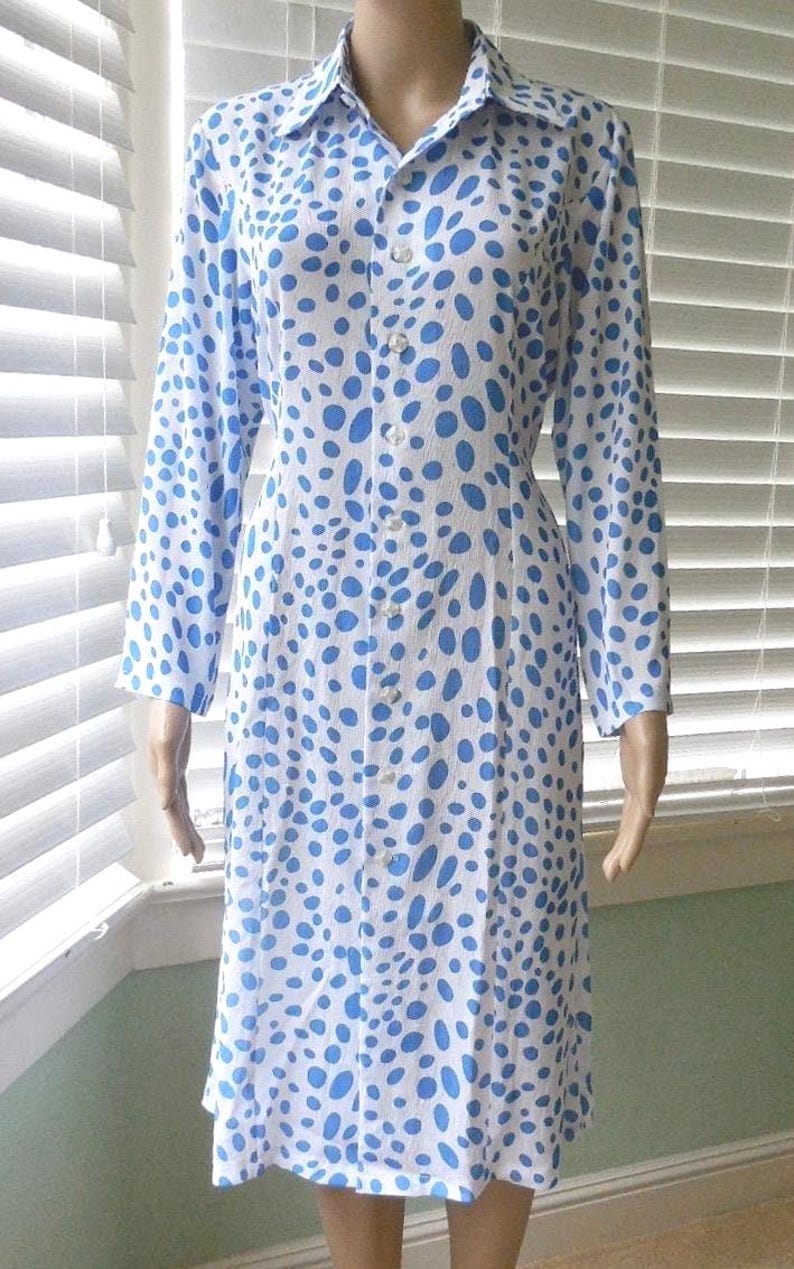 May include: A white dress with a blue polka dot pattern. The dress features long sleeves, a collared neckline, and a button-down front. The dress is knee-length and has a fitted shape.