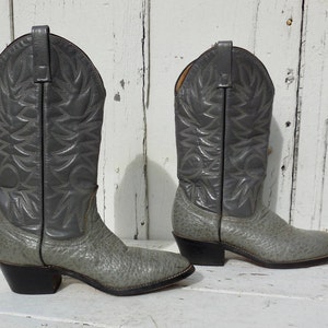 May include: A pair of gray cowboy boots with a textured finish. The boots have a classic design with pointed toes, decorative stitching, and black heels. The boots are made of leather and are suitable for various occasions.
