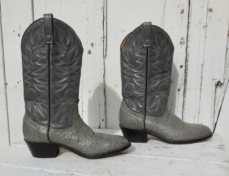May include: A pair of gray cowboy boots with a textured finish. The boots have a classic design with pointed toes, decorative stitching, and black heels. The boots are made of leather and are suitable for various occasions.