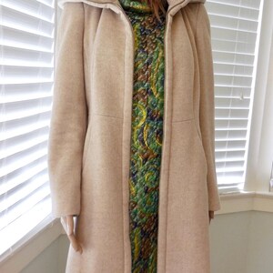 May include: A light beige coat with a high collar and a full-length zipper. The coat is worn over a patterned dress with green, blue, and gold tones. The mannequin is positioned in front of a window with white blinds.