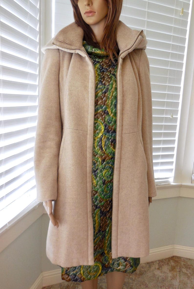 May include: A light beige coat with a high collar and a full-length zipper. The coat is worn over a patterned dress with green, blue, and gold tones. The mannequin is positioned in front of a window with white blinds.