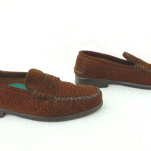 May include: A pair of brown loafers featuring a textured, pebbled surface across the upper. The shoes have a classic slip-on design with a stitched moccasin-style toe seam, a low heel, and a visible teal-colored insole inside the front shoe.