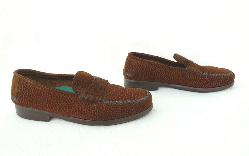 May include: A pair of brown loafers featuring a textured, pebbled surface across the upper. The shoes have a classic slip-on design with a stitched moccasin-style toe seam, a low heel, and a visible teal-colored insole inside the front shoe.