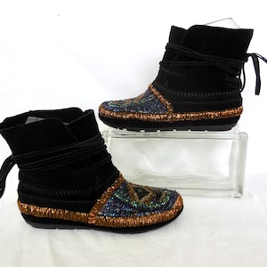 K&ouml;nnte beinhalten: Ein Paar schwarze Wildleder-Ankle Boots mit Perlenbesatz. Die Stiefel haben einen Wickelverschluss und sind mit farbigen Perlen in Blau-, Gr&uuml;n- und Goldt&ouml;nen verziert. Die Sohlen sind schwarz.