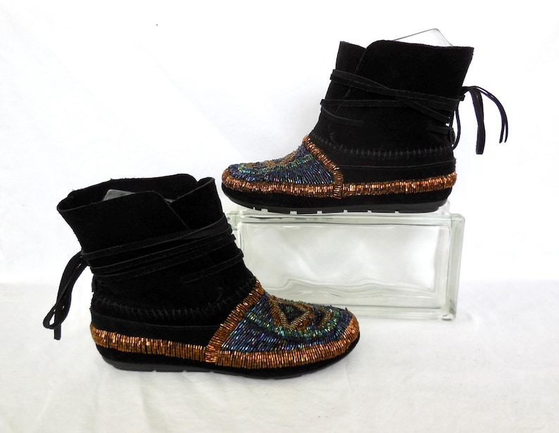 K&ouml;nnte beinhalten: Ein Paar schwarze Wildleder-Ankle Boots mit Perlenbesatz. Die Stiefel haben einen Wickelverschluss und sind mit farbigen Perlen in Blau-, Gr&uuml;n- und Goldt&ouml;nen verziert. Die Sohlen sind schwarz.