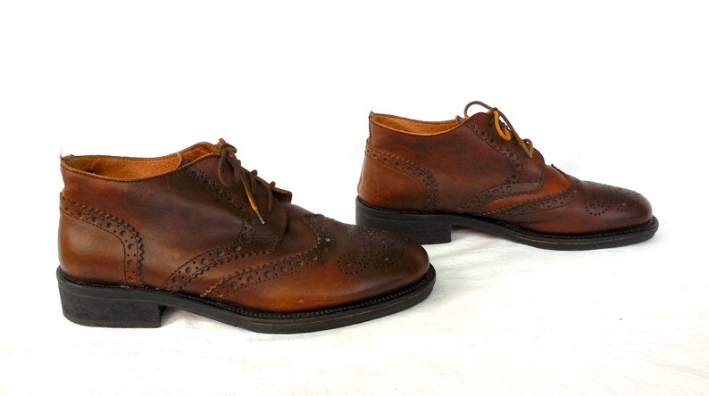 Pode incluir: Um par de botas de tornozelo de couro marrom com detalhes de brogue e fecho de cadar&ccedil;o.