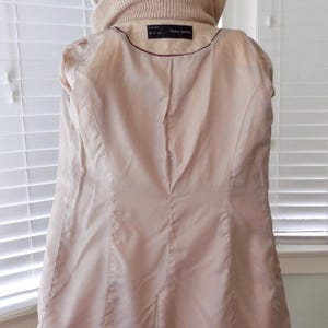 May include: A light beige coat with a hood and a ribbed collar. The inside lining is a light pink color. The coat has a tailored fit and a label that reads "ZARA BASIC".