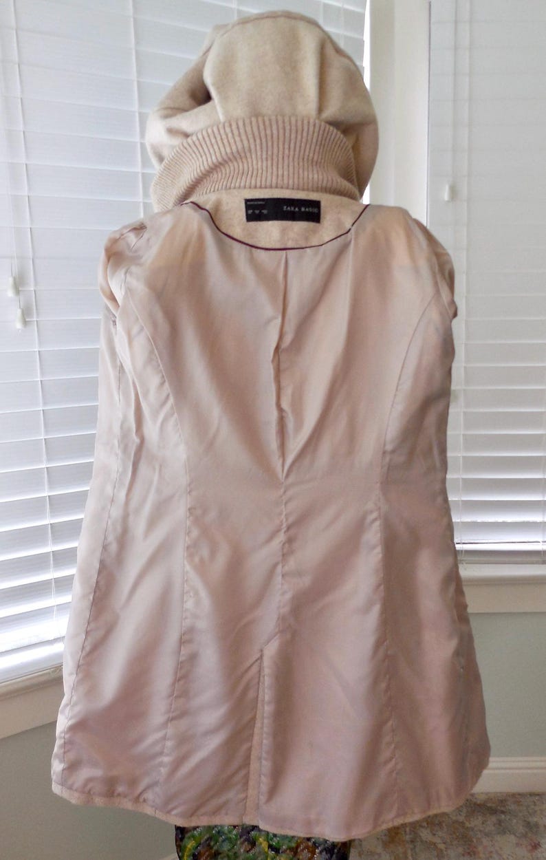 May include: A light beige coat with a hood and a ribbed collar. The inside lining is a light pink color. The coat has a tailored fit and a label that reads "ZARA BASIC".