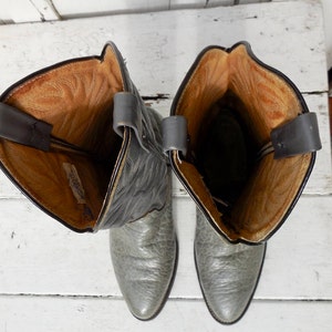 May include: A pair of gray cowboy boots with a textured leather appearance. The boots have a pointed toe and a tall shaft with pull straps. The interior is a light brown color.