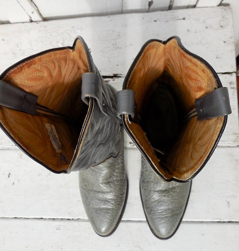 May include: A pair of gray cowboy boots with a textured leather appearance. The boots have a pointed toe and a tall shaft with pull straps. The interior is a light brown color.