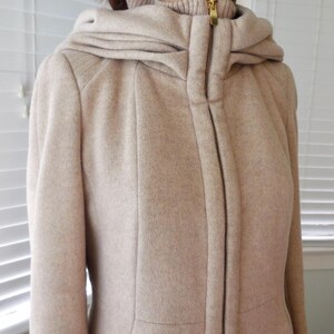May include: A beige hooded coat with a full-length zipper and a high collar. The coat has a tailored fit and a hood that drapes over the shoulders. The zipper is gold-toned. The coat is made of a textured fabric.