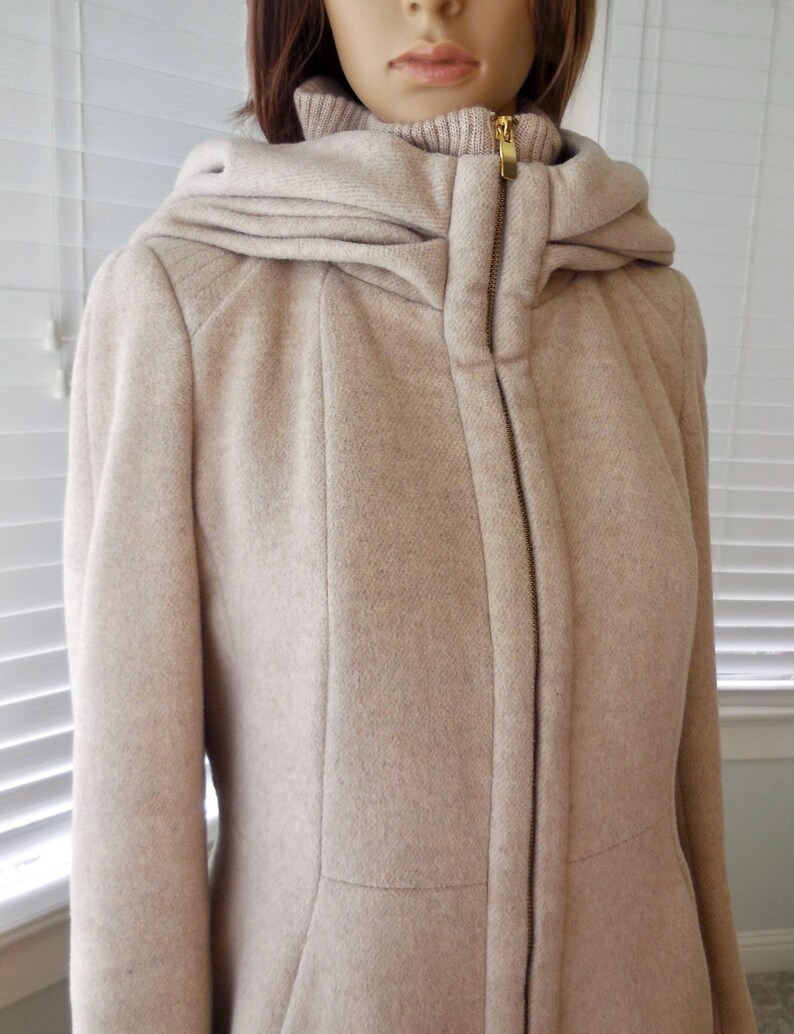May include: A beige hooded coat with a full-length zipper and a high collar. The coat has a tailored fit and a hood that drapes over the shoulders. The zipper is gold-toned. The coat is made of a textured fabric.