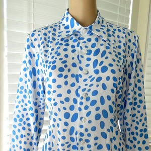 May include: A white shirt with a blue polka dot pattern. The long-sleeved shirt has a classic collar and is buttoned up the front. The blue dots vary in size and shape, creating a playful design. The shirt is made of a lightweight fabric.