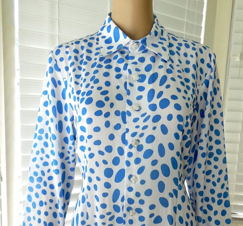 May include: A white shirt with a blue polka dot pattern. The long-sleeved shirt has a classic collar and is buttoned up the front. The blue dots vary in size and shape, creating a playful design. The shirt is made of a lightweight fabric.
