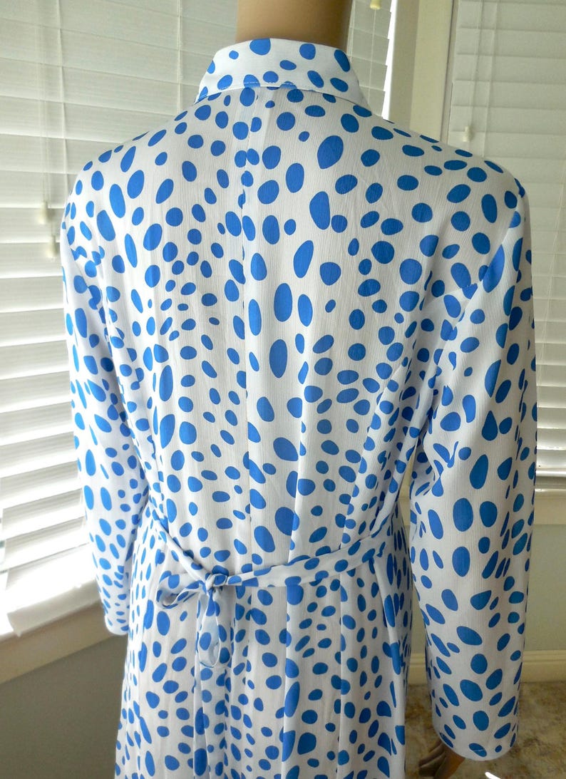 May include: White dress with a blue polka dot pattern. The dress has long sleeves, a collar, and a tie belt at the waist. The dress is made of a lightweight fabric.