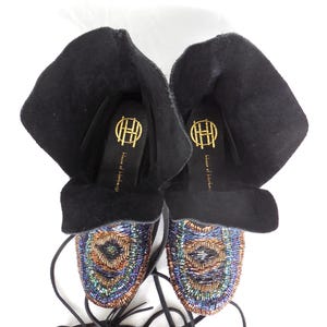 K&ouml;nnte beinhalten: Schwarze Wildleder-Mokassin-Stiefel mit aufwendiger Perlenarbeit in Blau, Gr&uuml;n und Kupfer. Die Stiefel haben einen umgeschlagenen Schaft und lange schwarze Schn&uuml;rsenkel. Die Innensohle zeigt ein goldenes Logo und den Text "House of Harlow 1960".