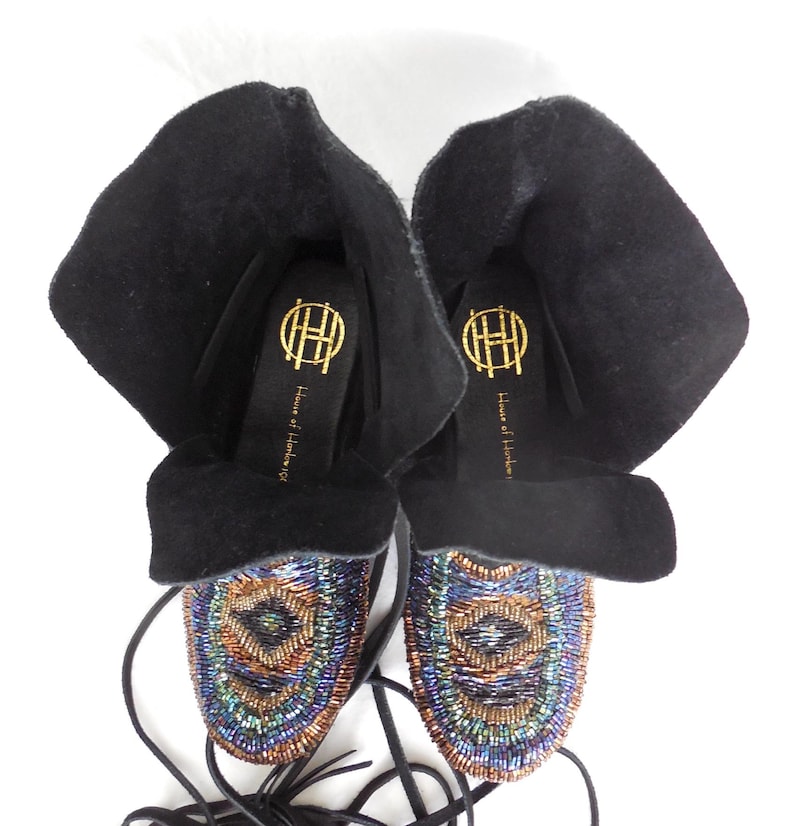 K&ouml;nnte beinhalten: Schwarze Wildleder-Mokassin-Stiefel mit aufwendiger Perlenarbeit in Blau, Gr&uuml;n und Kupfer. Die Stiefel haben einen umgeschlagenen Schaft und lange schwarze Schn&uuml;rsenkel. Die Innensohle zeigt ein goldenes Logo und den Text "House of Harlow 1960".