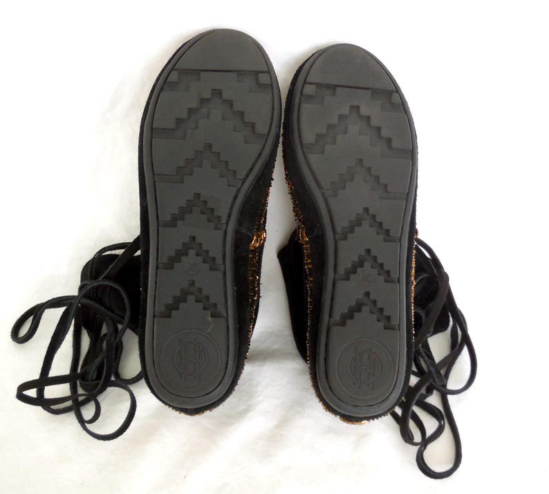 K&ouml;nnte beinhalten: Ein Paar schwarze Ballerinas mit goldenen Akzenten, von unten gesehen. Die schwarzen Sohlen haben ein geometrisches Profil. Lange schwarze Schn&uuml;rsenkel sind zu sehen. Auf wei&szlig;em Hintergrund.