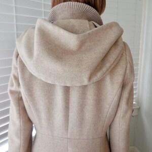 May include: A beige wool coat with a hood and a high collar. The coat has a tailored fit and a button closure. The hood is large and covers the shoulders. The coat is made of a soft, textured fabric.