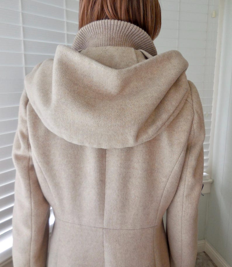 May include: A beige wool coat with a hood and a high collar. The coat has a tailored fit and a button closure. The hood is large and covers the shoulders. The coat is made of a soft, textured fabric.