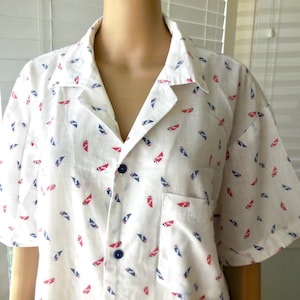 May include: White button-down shirt with a pattern of red and blue shapes. The short-sleeved shirt has a collar, a chest pocket, and blue buttons. The fabric appears to be lightweight.