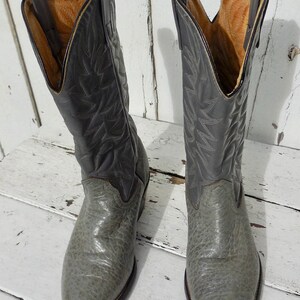 May include: A pair of gray cowboy boots with a textured finish. The boots have a pointed toe and a tall shaft with decorative stitching. The interior is a light brown color. The boots are made of leather and are likely vintage.