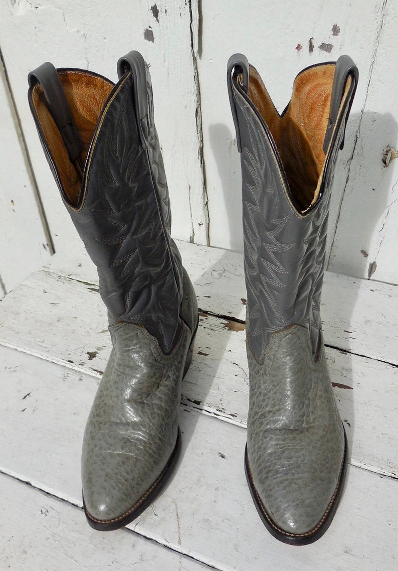 May include: A pair of gray cowboy boots with a textured finish. The boots have a pointed toe and a tall shaft with decorative stitching. The interior is a light brown color. The boots are made of leather and are likely vintage.