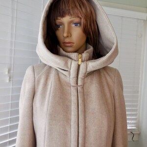 May include: A beige hooded coat with a gold zipper, displayed on a mannequin. The coat has a high collar and a matching hood. The fabric appears to be a textured wool blend, offering a warm and stylish outerwear option.