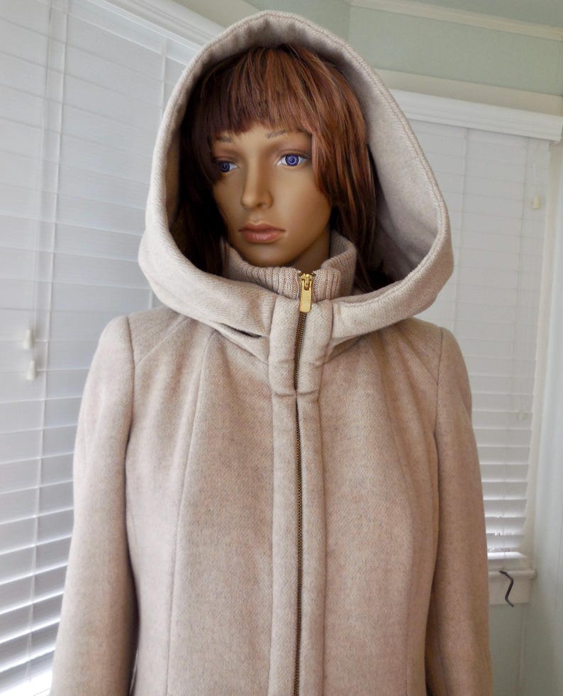 May include: A beige hooded coat with a gold zipper, displayed on a mannequin. The coat has a high collar and a matching hood. The fabric appears to be a textured wool blend, offering a warm and stylish outerwear option.