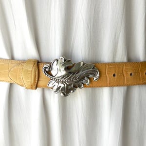 May include: A tan leather belt with a silver leaf-shaped buckle. The belt has a crocodile texture and is fastened around a white garment. The buckle is detailed with intricate leaf veins and a beaded border.