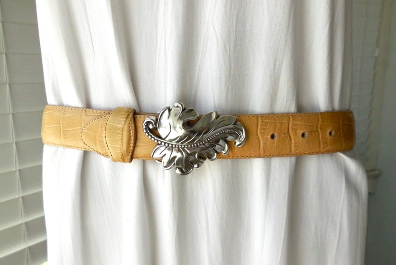 May include: A tan leather belt with a silver leaf-shaped buckle. The belt has a crocodile texture and is fastened around a white garment. The buckle is detailed with intricate leaf veins and a beaded border.