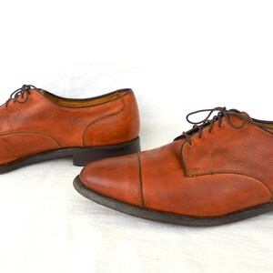 May include: A pair of brown leather dress shoes with dark brown laces. The shoes have a classic cap-toe design and a dark sole. The leather appears worn, giving the shoes a vintage look. The shoes are displayed on a white background.