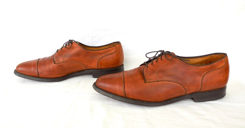 May include: A pair of brown leather dress shoes with dark brown laces. The shoes have a classic cap-toe design and a dark sole. The leather appears worn, giving the shoes a vintage look. The shoes are displayed on a white background.