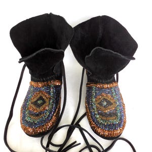 Peut inclure: Paire de bottes mocassins en daim noir avec des détails perlés. Les bottes ont un revers et de longs lacets noirs. Le motif perlé présente un motif circulaire avec des nuances de bleu, de vert et de cuivre. Chaussures uniques et élégantes.