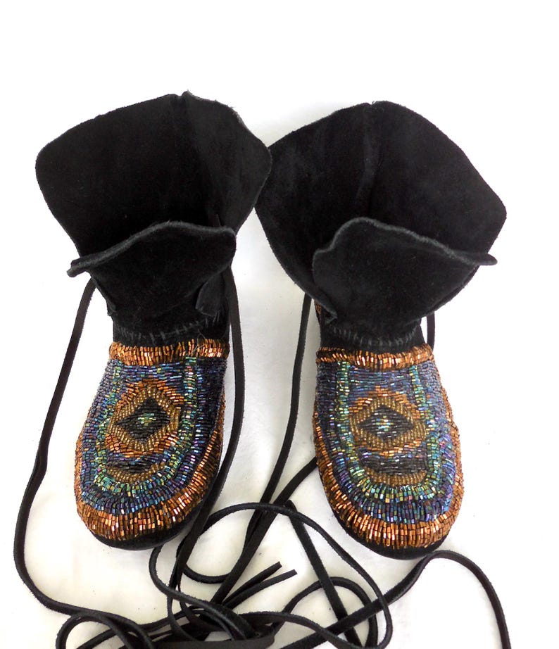 K&ouml;nnte beinhalten: Ein Paar schwarze Wildleder-Mokassin-Stiefel mit Perlenverzierungen. Die Stiefel haben einen umklappbaren Schaft und lange schwarze Schn&uuml;rsenkel. Das Perlenmuster zeigt ein kreisf&ouml;rmiges Muster mit Blau-, Gr&uuml;n- und Kupfert&ouml;nen. Einzigartige und stilvolle Schuhe.