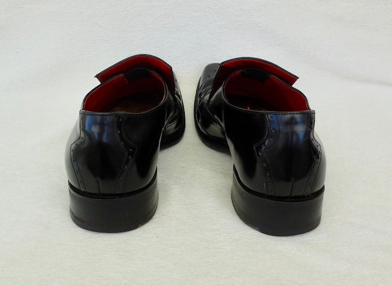 Puede incluir: Un par de zapatos de vestir de cuero negro con dise&ntilde;o de punta de ala. Los zapatos tienen un forro de cuero rojo y una suela de goma negra.