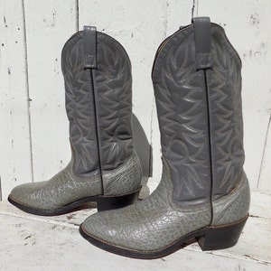 May include: A pair of gray cowboy boots. The boots have a textured, light gray foot and a smooth, darker gray shaft with decorative stitching. The heels are black, and the boots are set against a white, weathered wooden background.