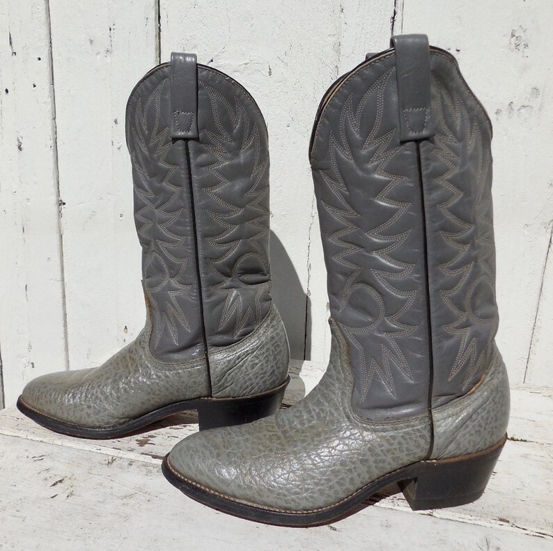 May include: A pair of gray cowboy boots. The boots have a textured, light gray foot and a smooth, darker gray shaft with decorative stitching. The heels are black, and the boots are set against a white, weathered wooden background.