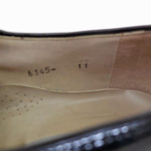 Puede incluir: Vista interior de un zapato negro, que muestra la plantilla y el interior del zapato. La plantilla es de color beige con peque&ntilde;os orificios de ventilaci&oacute;n. El n&uacute;mero "11" est&aacute; impreso cerca del tal&oacute;n y el n&uacute;mero "6345-" cerca de la puntera.