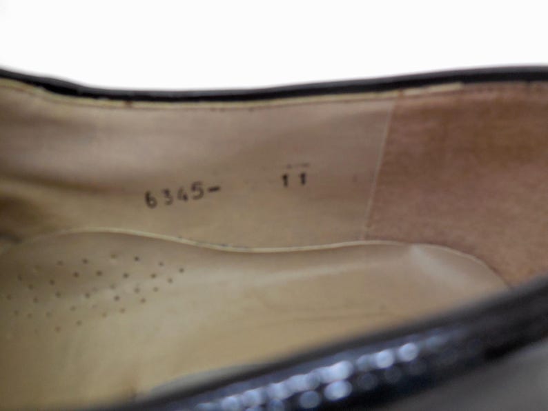 Puede incluir: Vista interior de un zapato negro, que muestra la plantilla y el interior del zapato. La plantilla es de color beige con peque&ntilde;os orificios de ventilaci&oacute;n. El n&uacute;mero "11" est&aacute; impreso cerca del tal&oacute;n y el n&uacute;mero "6345-" cerca de la puntera.