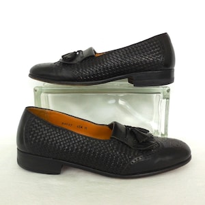May include: A pair of black leather tassel loafers with a woven pattern. The loafers have a brogue design and a rounded toe.