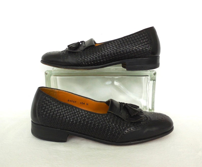 May include: A pair of black leather tassel loafers with a woven pattern. The loafers have a brogue design and a rounded toe.