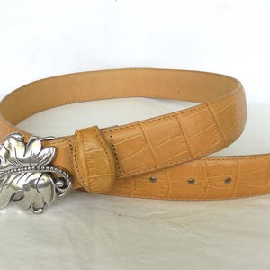 May include: Tan leather belt with a silver leaf-shaped buckle. The belt has a textured, crocodile-like pattern. The buckle is detailed with a beaded edge. A stylish accessory for any outfit.
