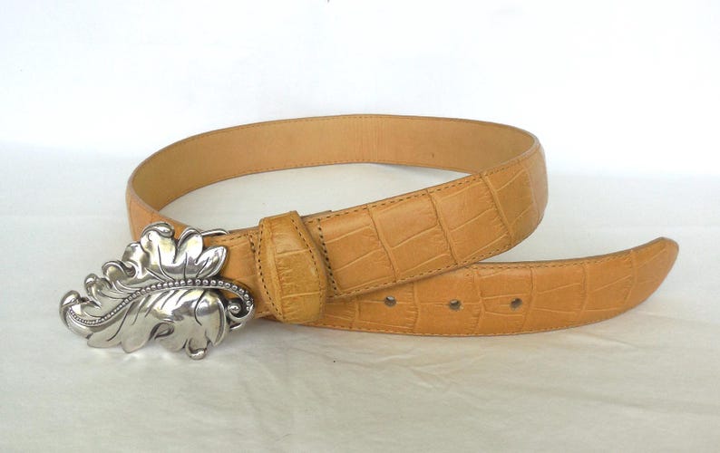 May include: Tan leather belt with a silver leaf-shaped buckle. The belt has a textured, crocodile-like pattern. The buckle is detailed with a beaded edge. A stylish accessory for any outfit.
