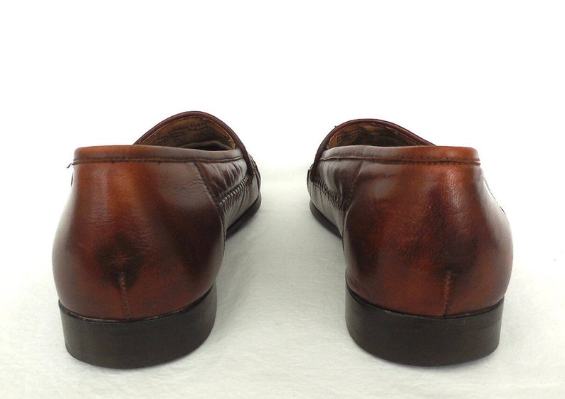 K&ouml;nnte beinhalten: Ein Paar braune Leder-Slipper mit einem gewebten Muster auf dem Spann. Die Slipper haben eine abgerundete Spitze und einen niedrigen Absatz.