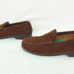 May include: A pair of brown loafers featuring a textured, pebbled surface finish. These slip-on shoes have a classic moccasin-style construction with a stitched apron toe, a low heel, and a dark brown sole. The interior lining is tan.