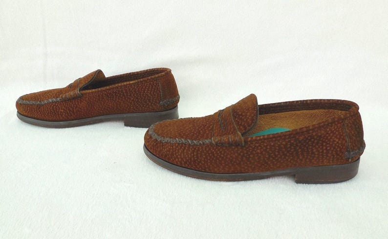 May include: A pair of brown loafers featuring a textured, pebbled surface finish. These slip-on shoes have a classic moccasin-style construction with a stitched apron toe, a low heel, and a dark brown sole. The interior lining is tan.
