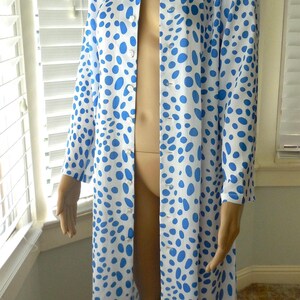 May include: A long, button-down robe featuring a white base and blue polka dots. The robe has long sleeves and a collar. The garment is displayed on a mannequin.