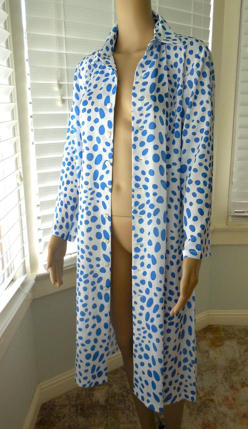 May include: A long, button-down robe featuring a white base and blue polka dots. The robe has long sleeves and a collar. The garment is displayed on a mannequin.